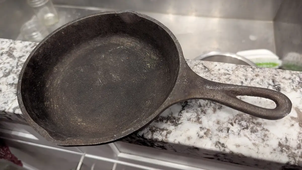 A well-used, dark, seasoned cast iron skillet rests on a speckled granite countertop near a stainless steel sink. The pan…
