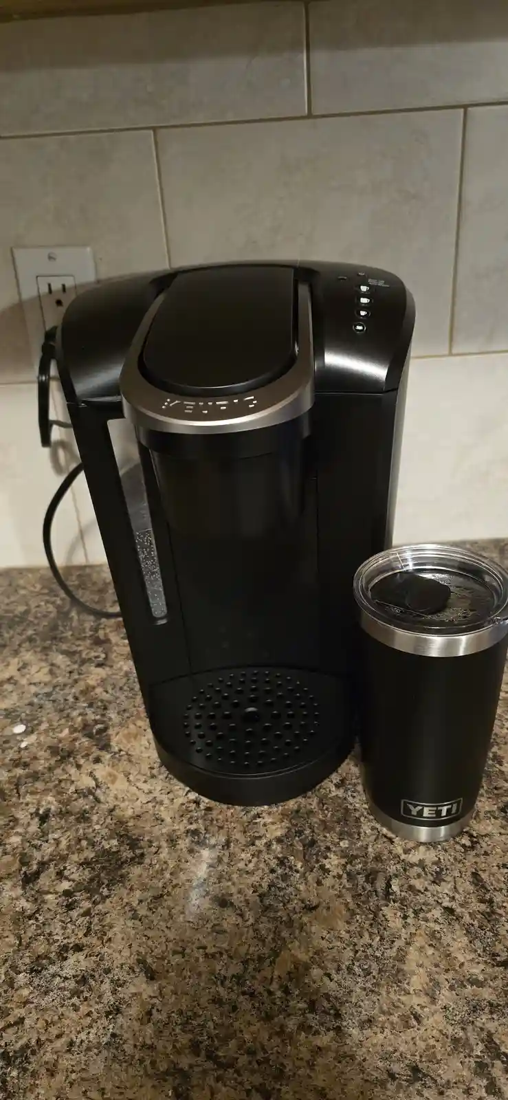 A black Keurig single-serve coffee maker sits on a speckled granite countertop next to a black Yeti insulated tumbler with a…