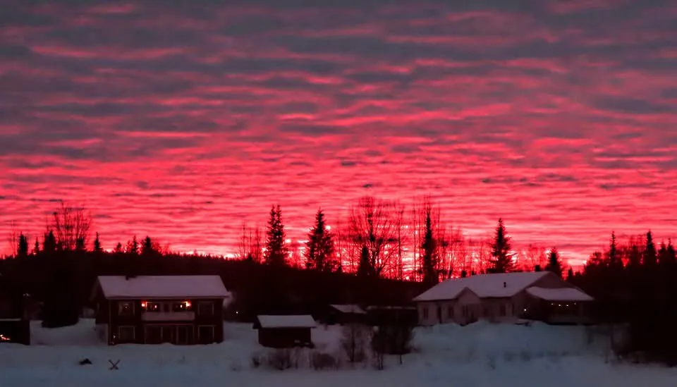 A snowy landscape at dusk or dawn with two houses and trees silhouetted against a sky filled with vivid, fiery red clouds…