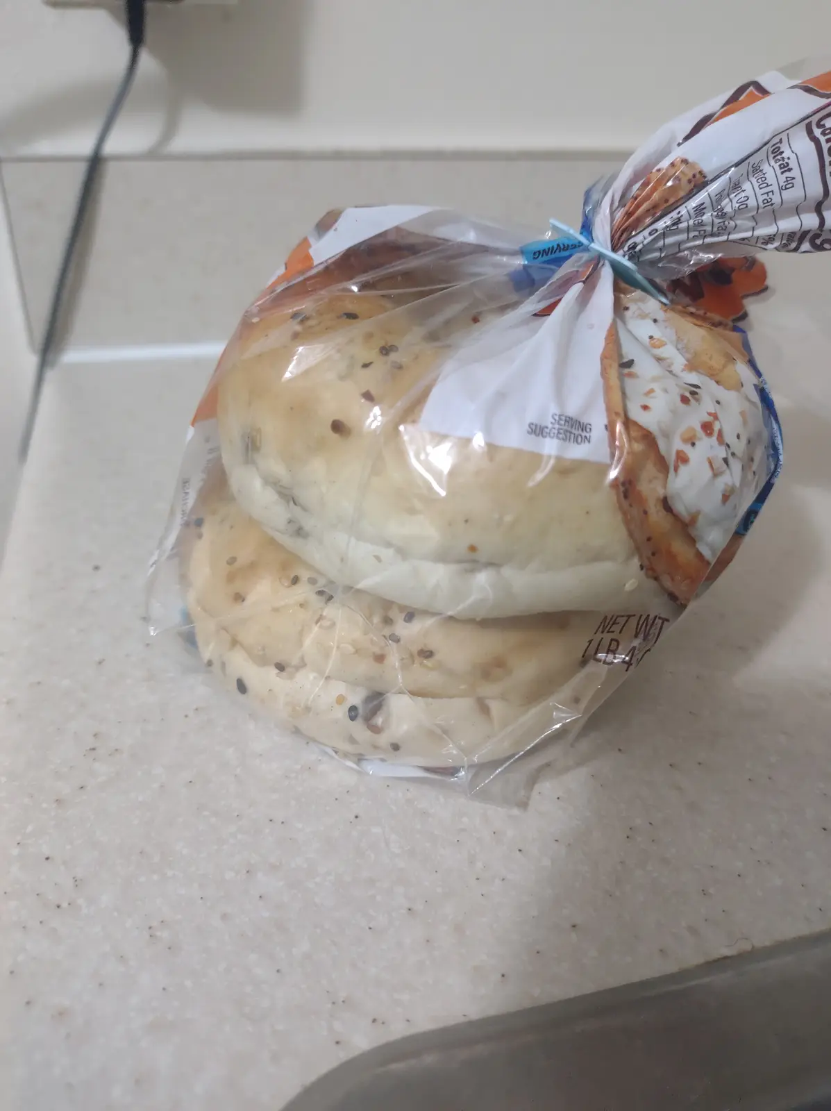 A plastic-wrapped bag of two round, seeded bread rolls sits on a speckled kitchen countertop. The bag is tied with a blue…