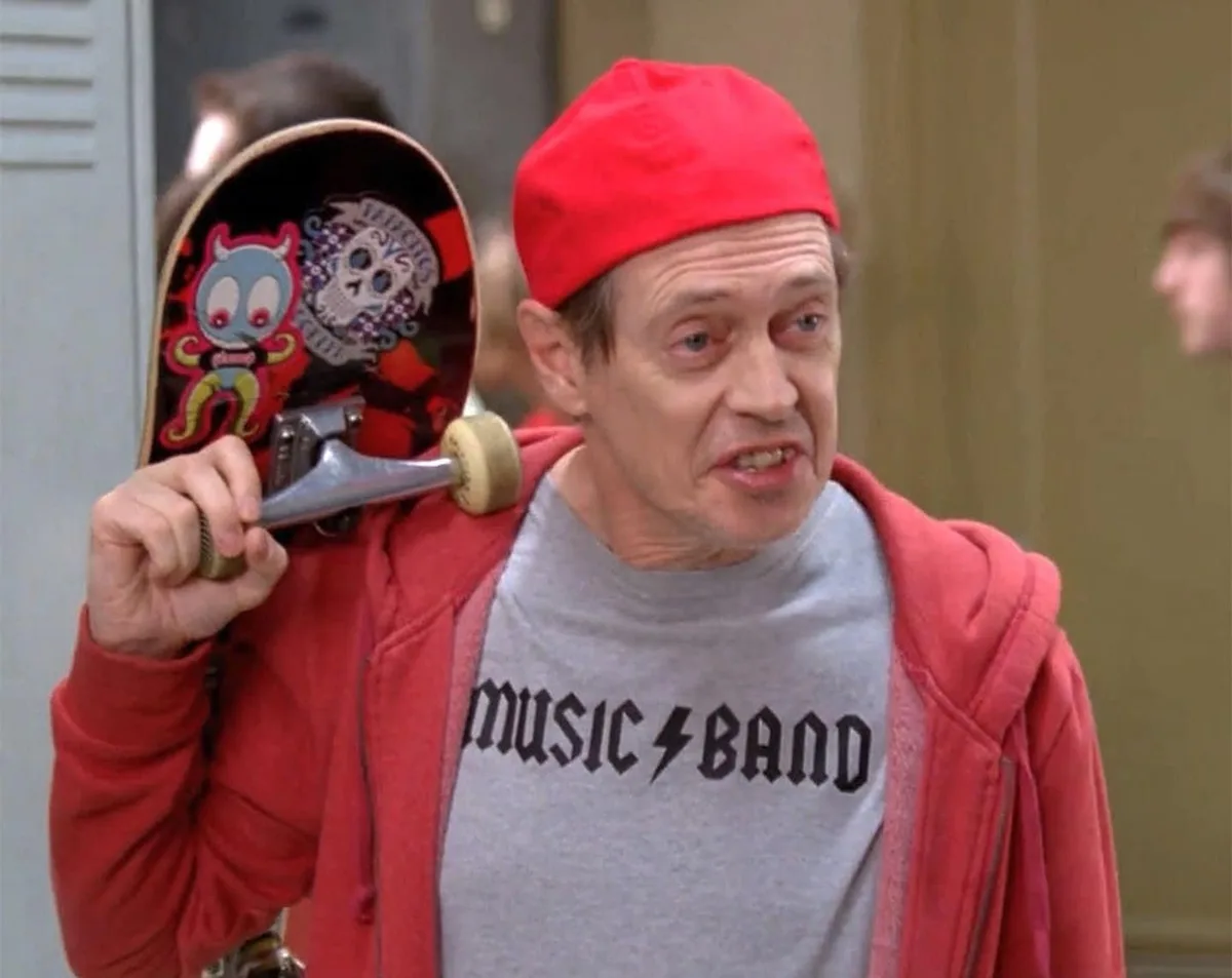A man wearing a red beanie, red hoodie, and a grey t-shirt with 'music band' printed on it holds a skateboard under his arm…