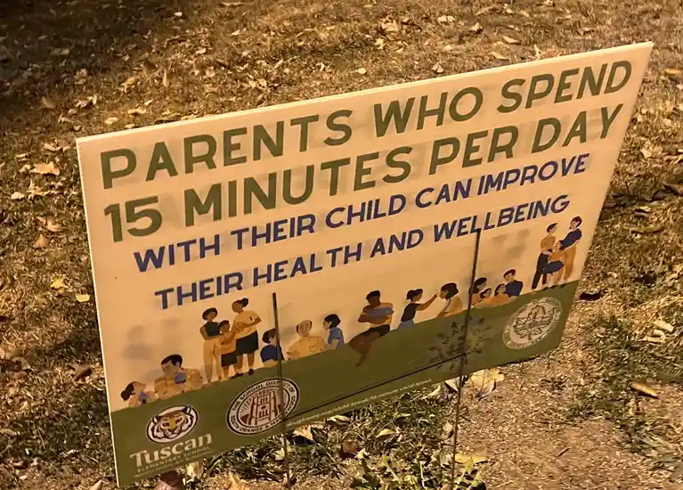 A white yard sign with green and blue text stands in grass at night, promoting parental engagement. The sign reads 'PARENTS…