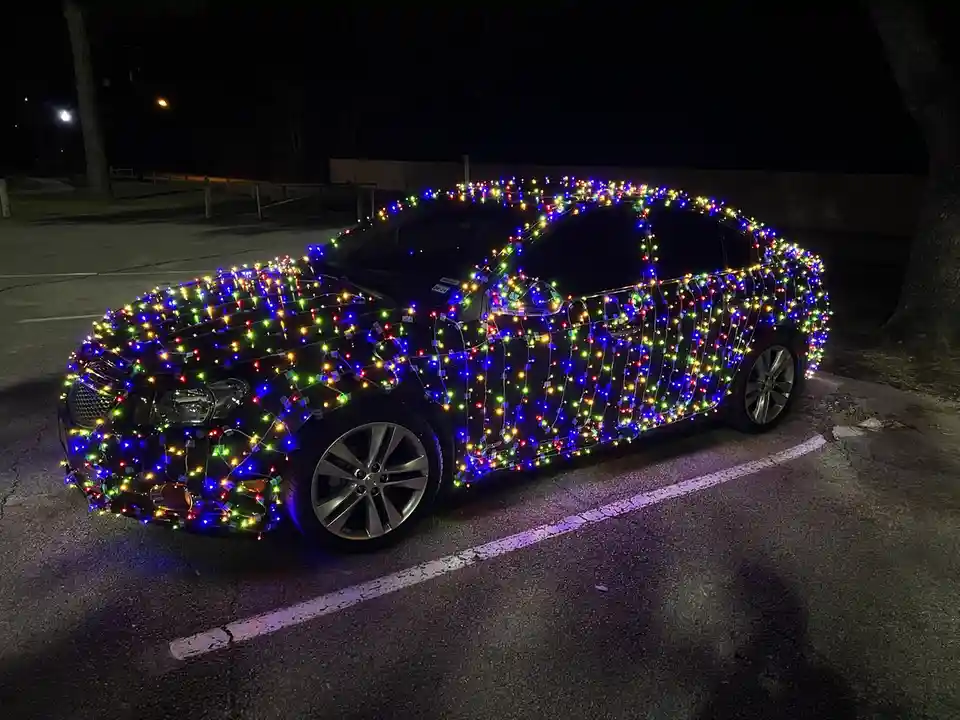 A black sedan parked at night is completely covered in multicolored string lights, resembling a glowing Christmas tree. The…