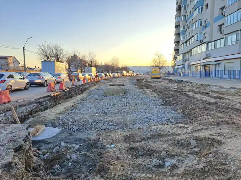 A street is dug up for construction with gravel and dirt, cars are parked on one side, and a yellow roller is visible in the…