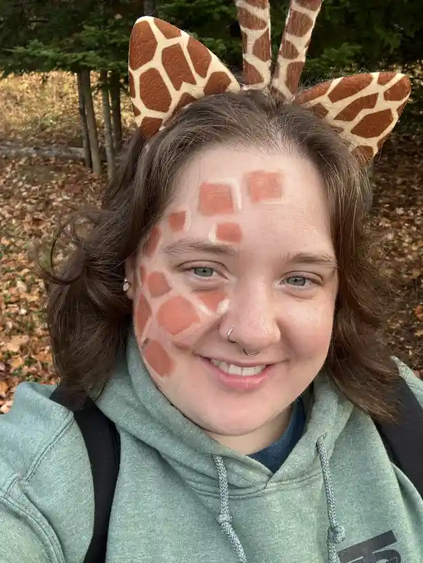 A person with brown hair and a nose ring smiles while wearing a giraffe-patterned headband with ears and horns, and has…