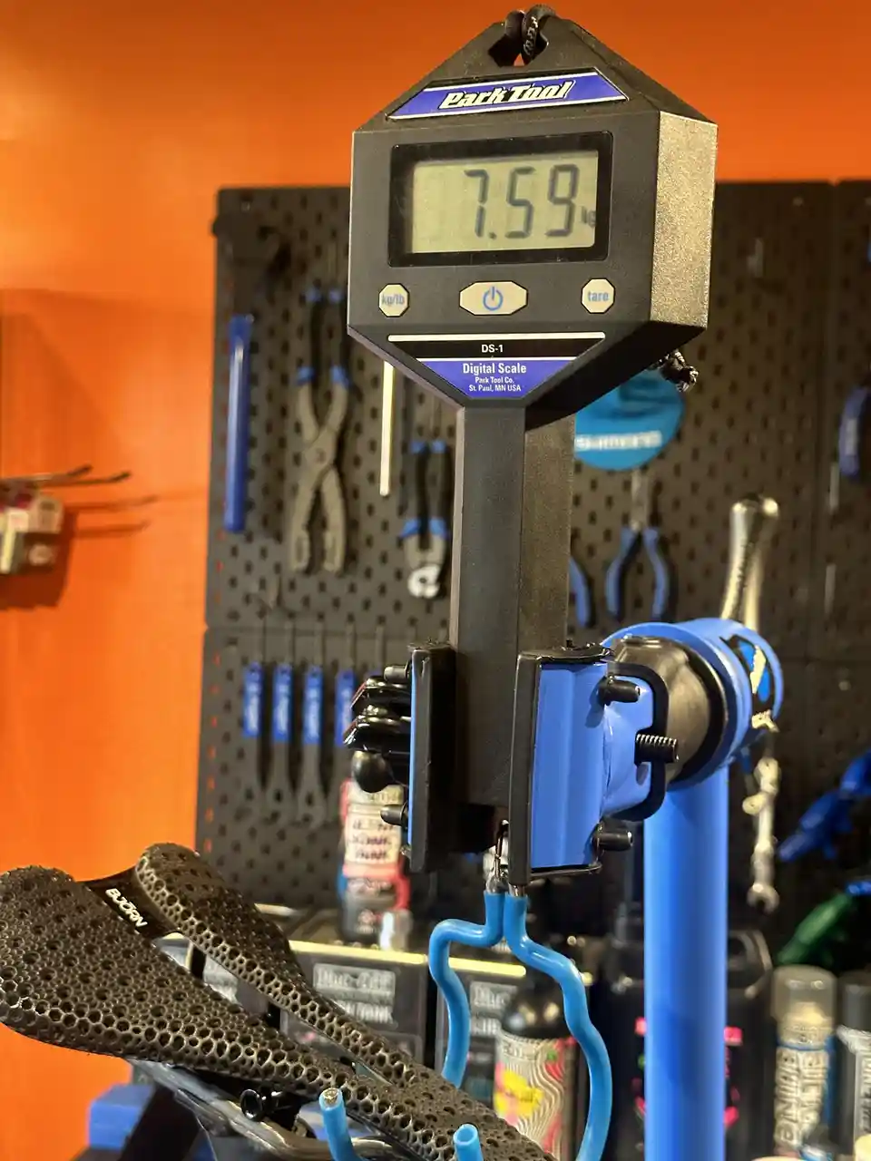A digital Park Tool scale displays '7.59 kg' while weighing a bicycle saddle mounted on a blue bike repair stand, with…