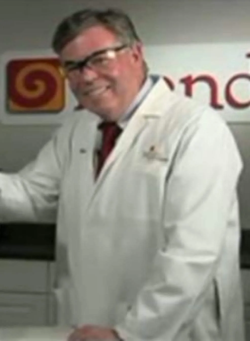 a man in a white lab coat and glasses smiles broadly while standing in front of a wall with a red spiral logo and partial…