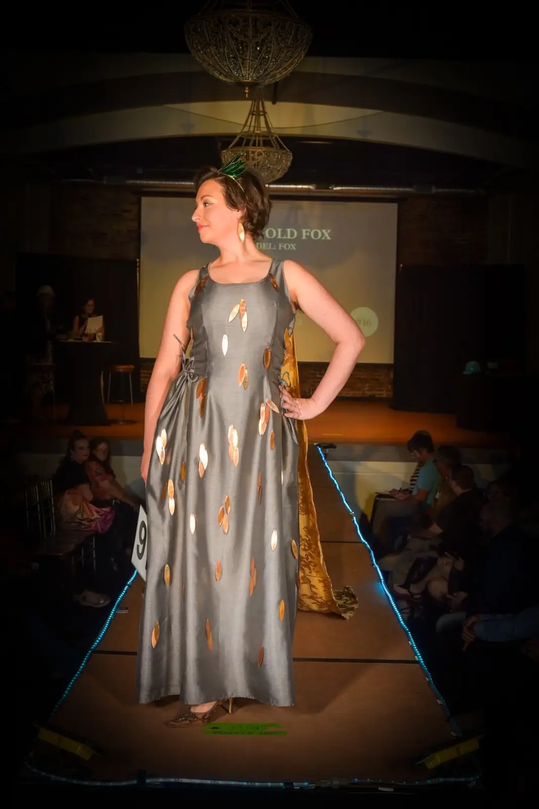 A woman walks down a runway in a grey dress adorned with gold leaf-like patterns, wearing a green headband and holding a…