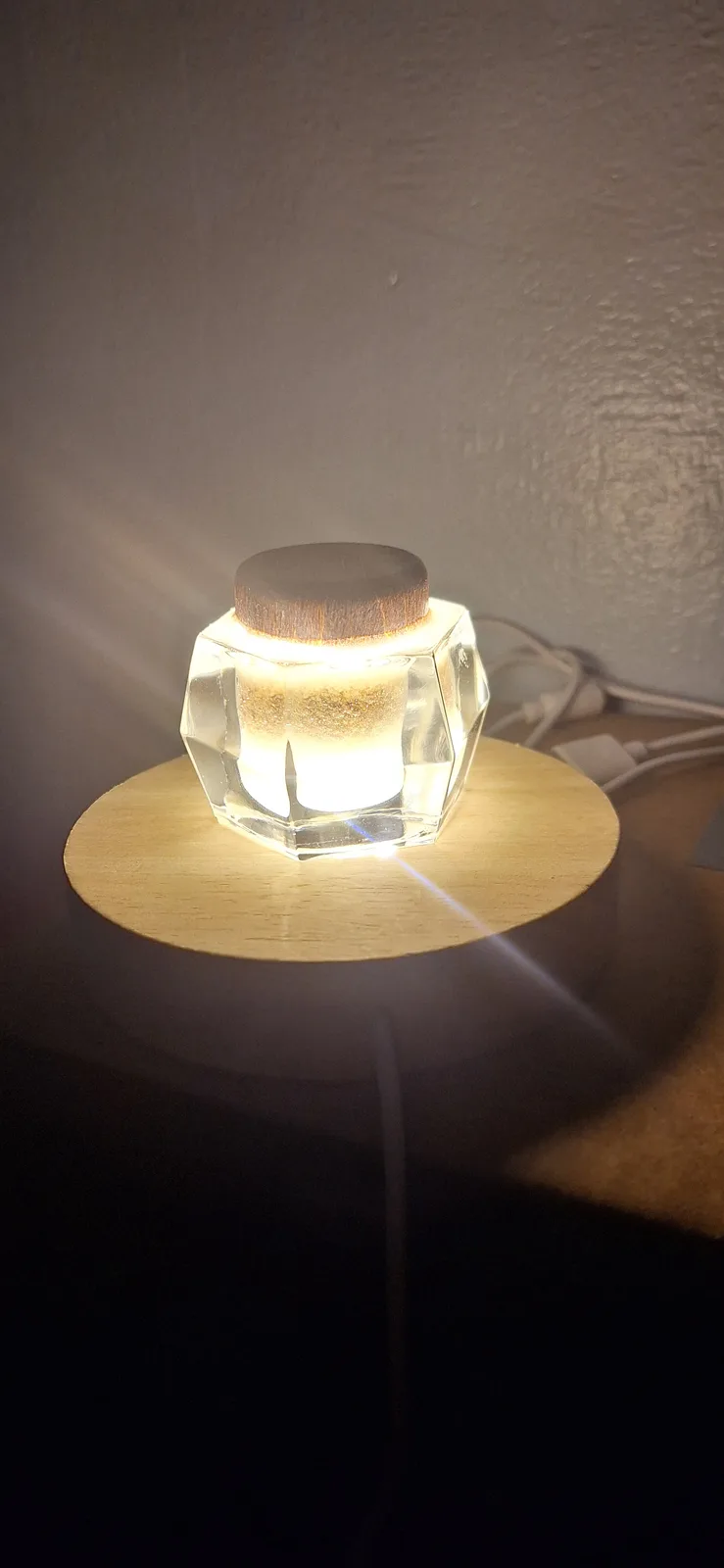 A close-up photo shows a glowing, geometric glass lamp with a wooden top, sitting on a circular wooden base. The lamp emits…
