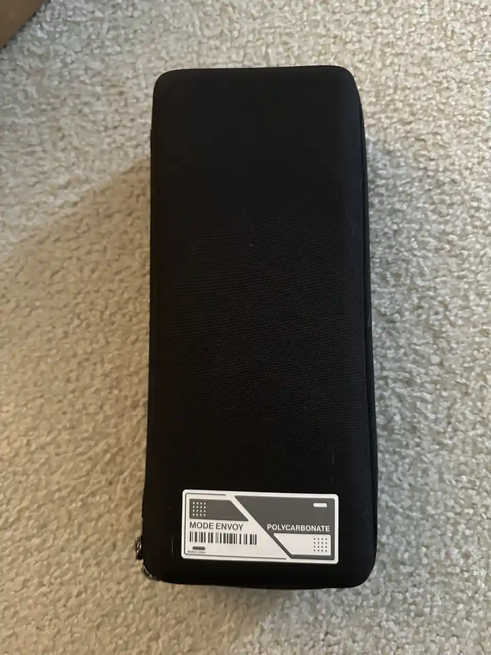 A black, textured, zippered case lies on a beige carpet, featuring a white label that reads 'MODE ENVOY POLYCARBONATE' with…