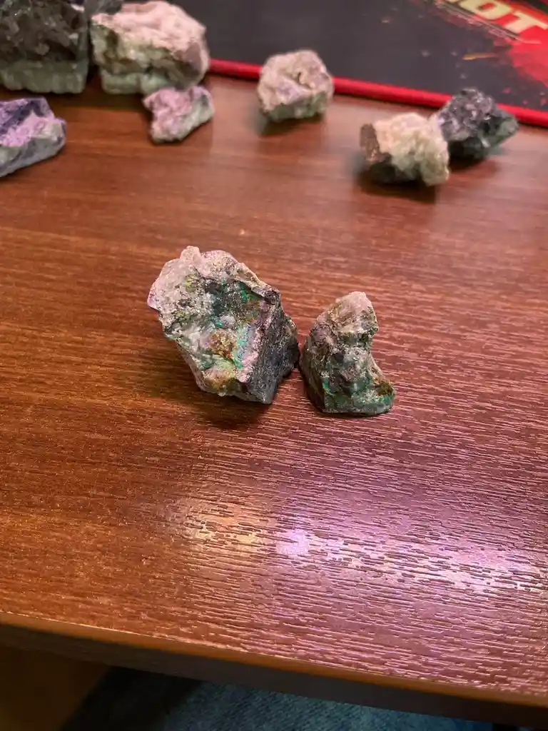 A close-up photo shows two small, rough, multi-colored mineral specimens sitting on a wooden table, with several other…