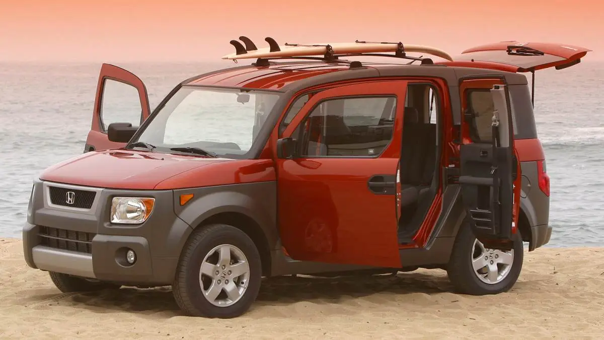 A red and gray Honda Element SUV is parked on a sandy beach with the ocean in the background. All four doors are open, and…