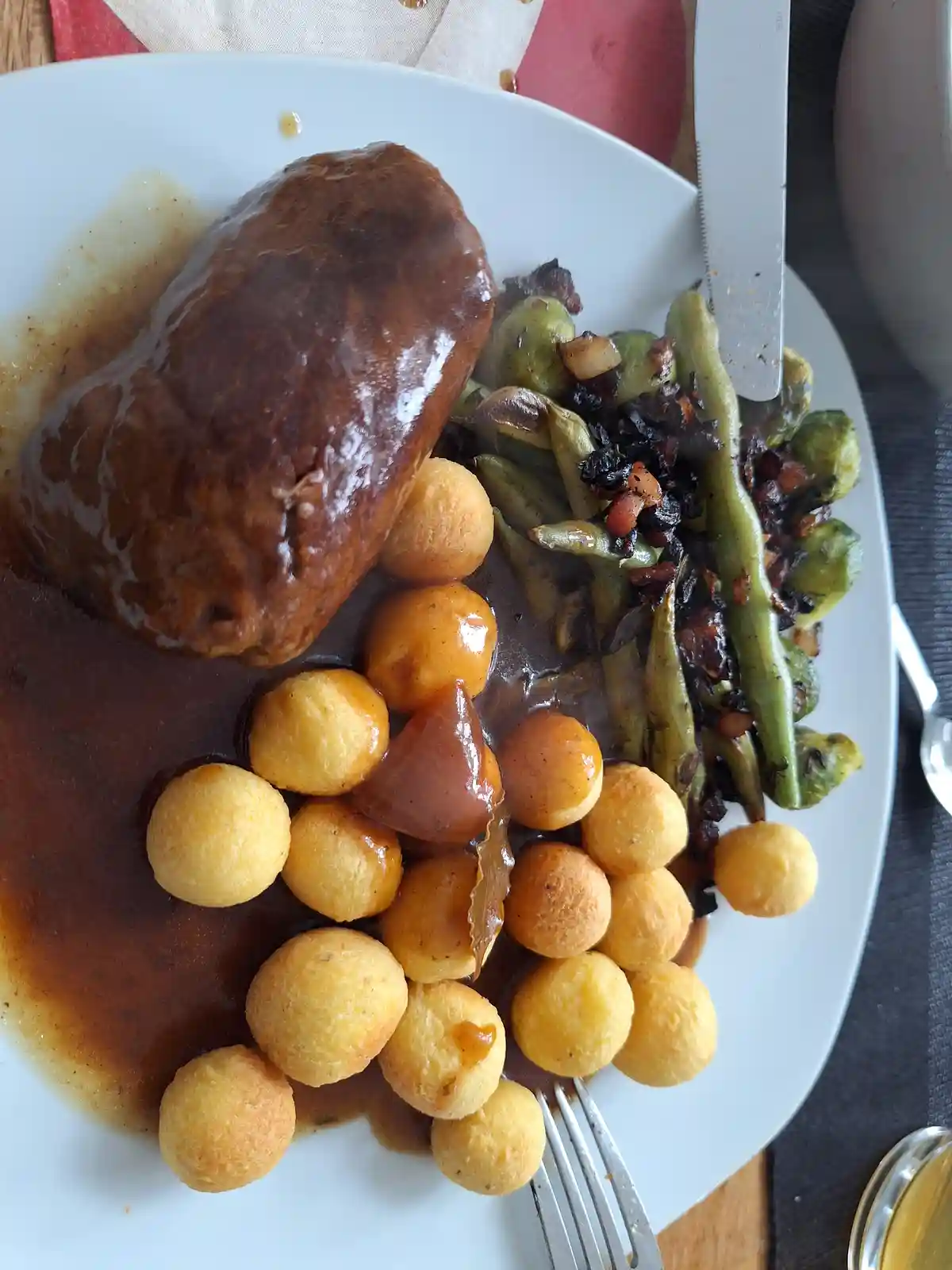 A close-up photo of a plated meal featuring a large, saucy meatloaf, golden-brown potato dumplings, and roasted green beans…