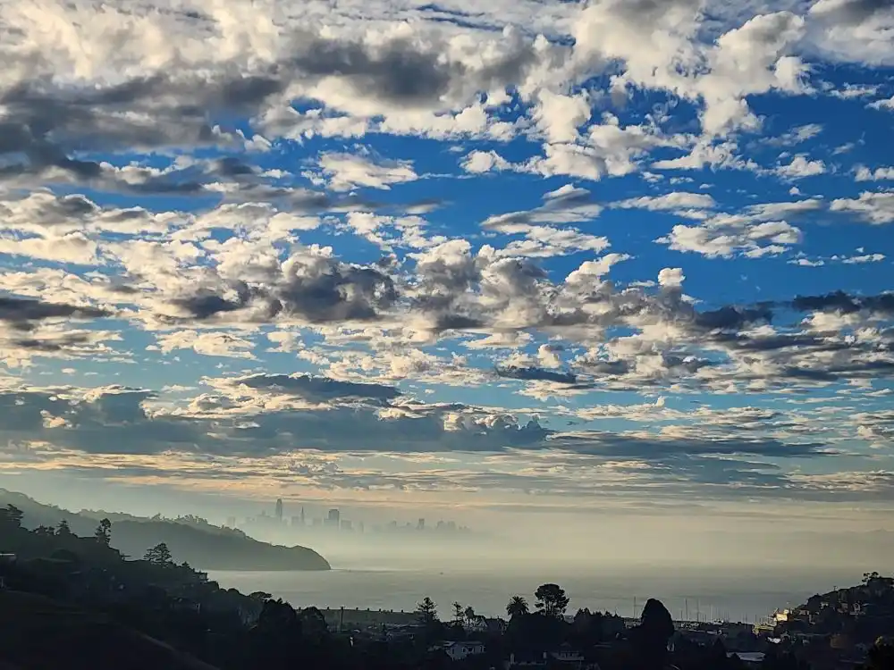 A scenic photo of a hazy coastal city skyline under a dramatic sky filled with scattered white and gray clouds against a…