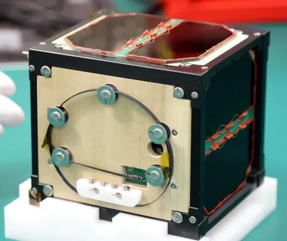 a close-up photo of a small, cube-shaped satellite with exposed circuitry, wires, and a solar panel on top, held by a gloved…