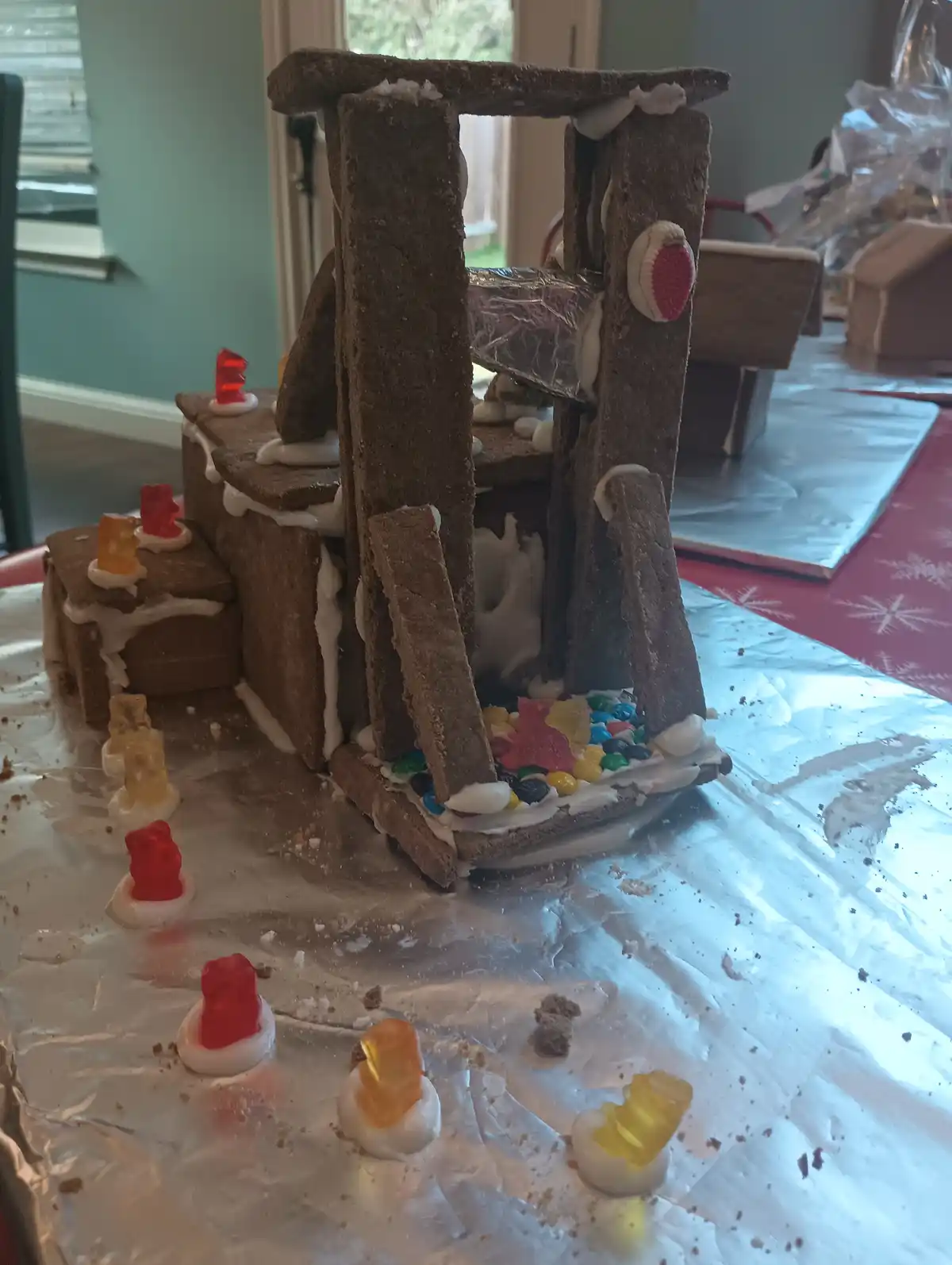 A gingerbread house made of brown graham crackers and white icing is visibly collapsing, with structural pieces leaning and…