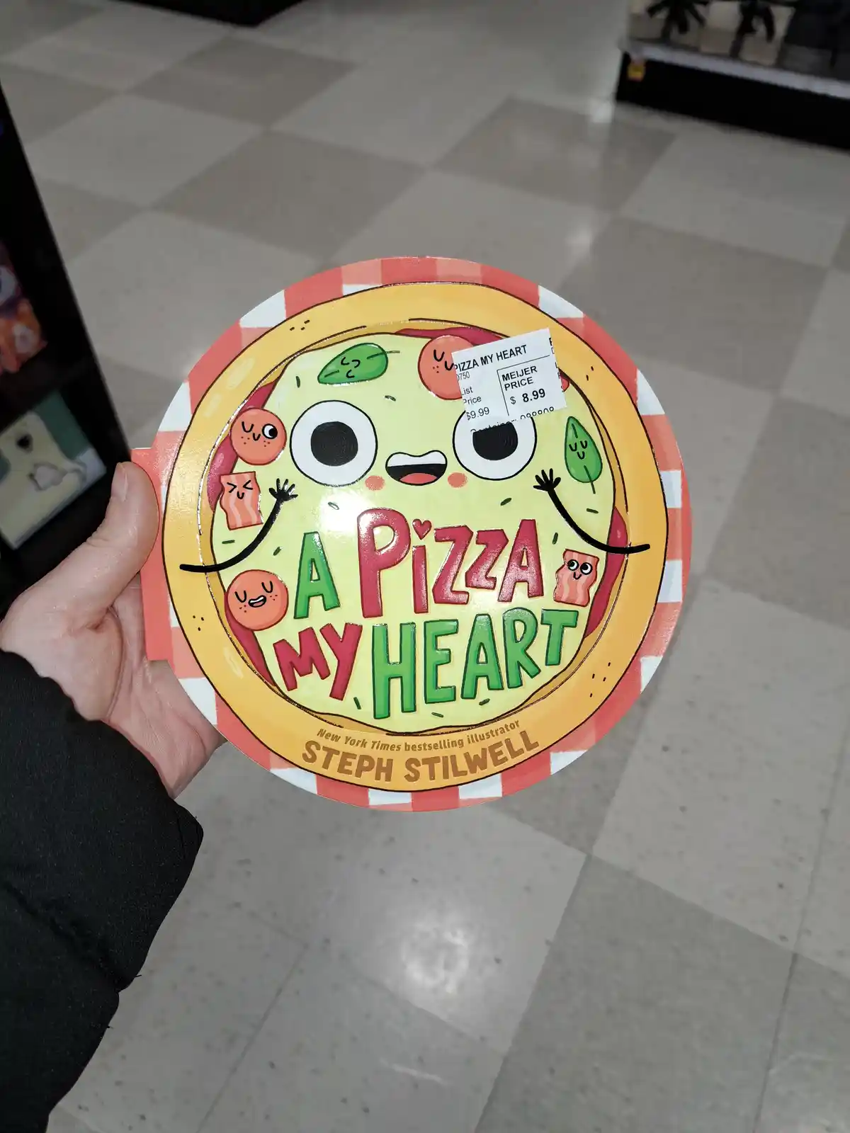 A hand holds a children's book titled 'A Pizza My Heart' by Steph Stilwell, featuring a cartoon pizza with smiling toppings…