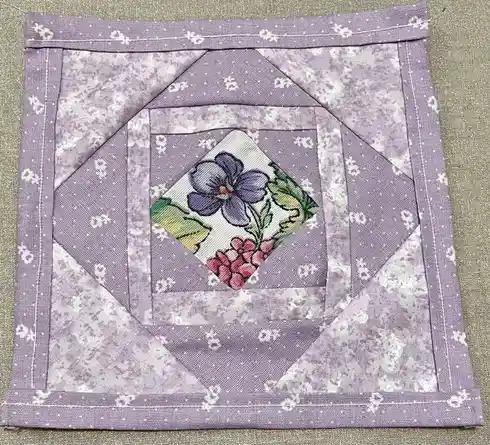 A handmade quilt block with a lavender and white patterned fabric, featuring a central diamond-shaped appliqué of a purple…