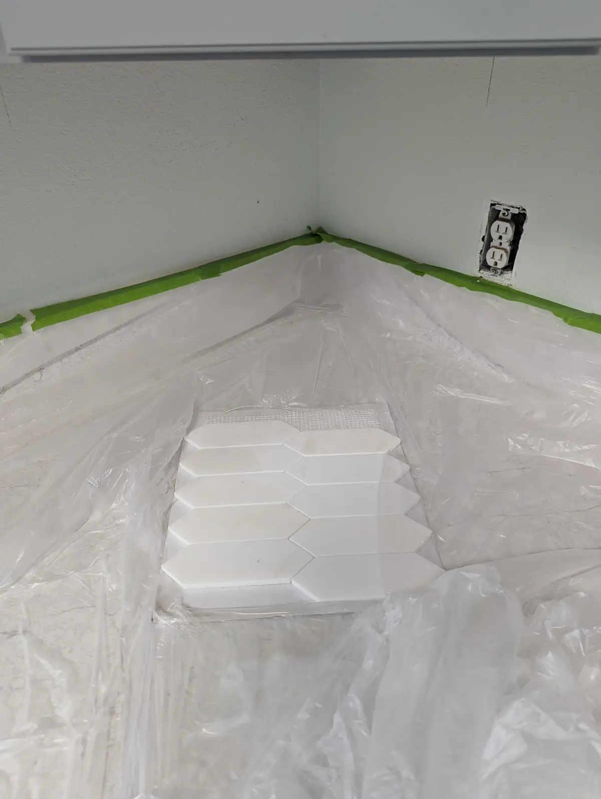 A close-up shot shows a corner of a room with white walls, where green painter's tape lines the baseboards and a white…