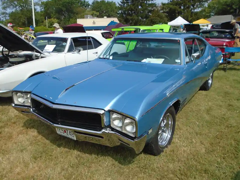 A shiny blue classic Buick Riviera is parked on grass at a car show, with other vintage cars and people in the background…
