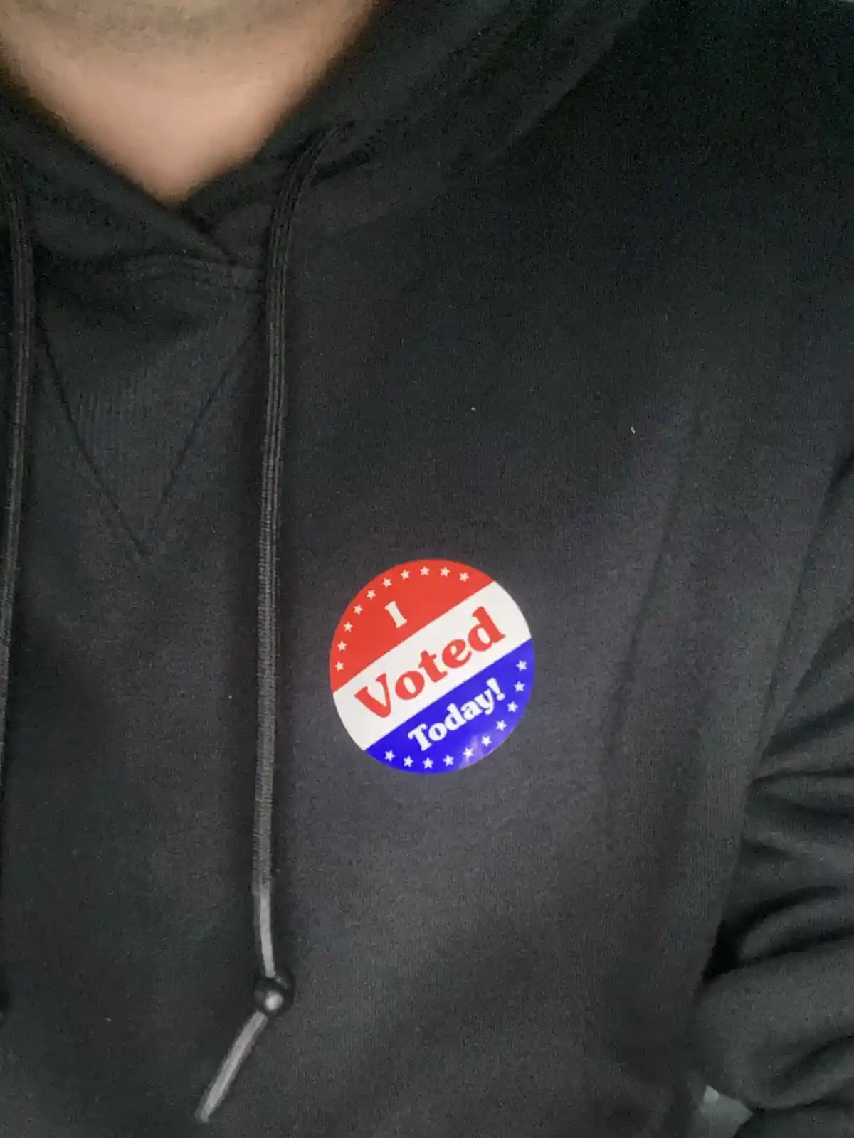 A close-up photo shows a person wearing a black hoodie with a circular red, white, and blue 'I Voted Today!' sticker pinned…