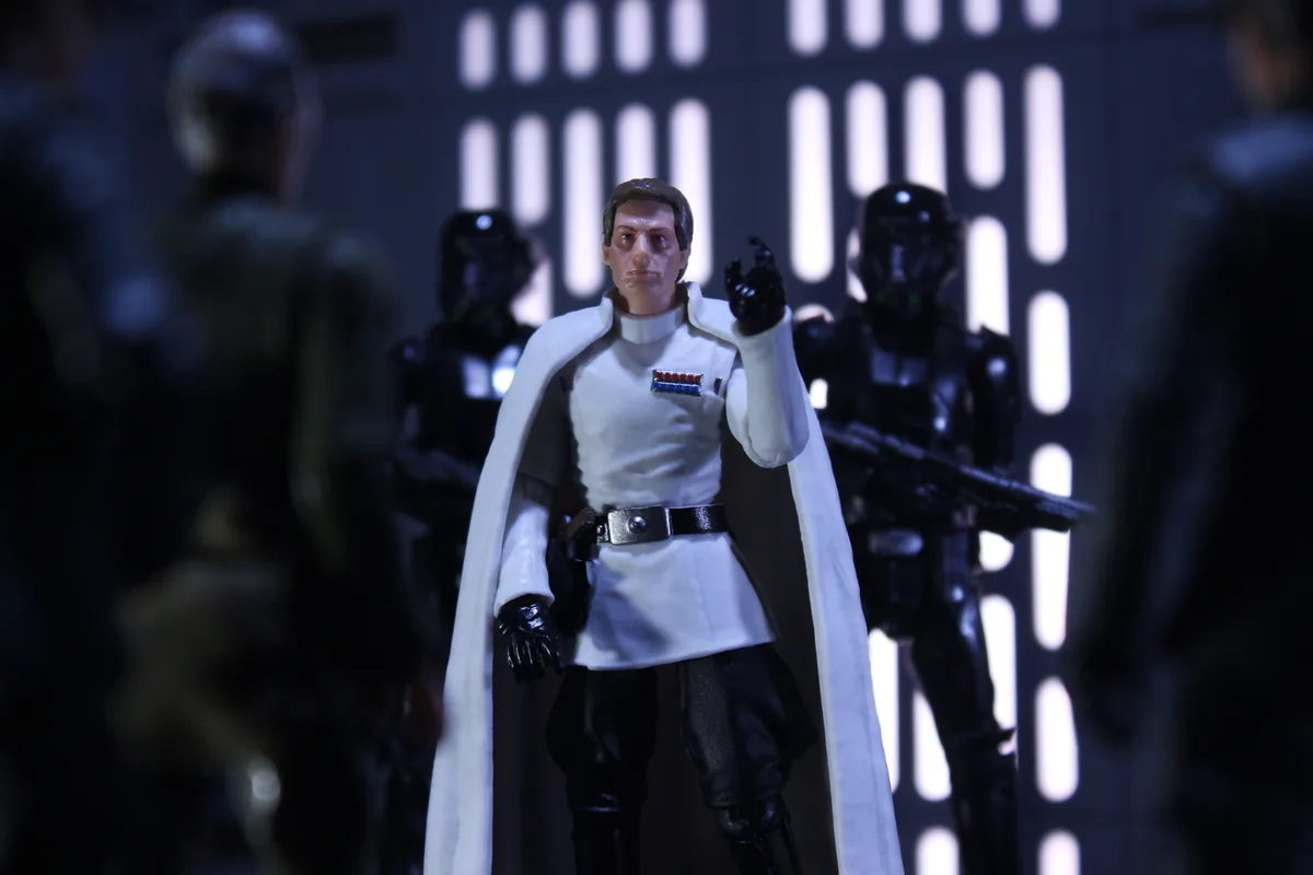 A Star Wars action figure of a white-clad Imperial officer stands in the center, pointing forward with a stern expression…