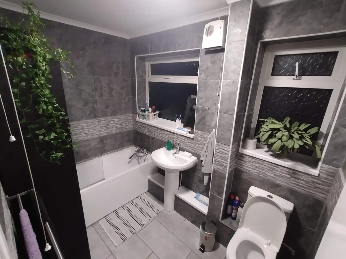 A modern bathroom with grey tiled walls, a white bathtub, sink, and toilet, featuring two windows with plants and a visible…