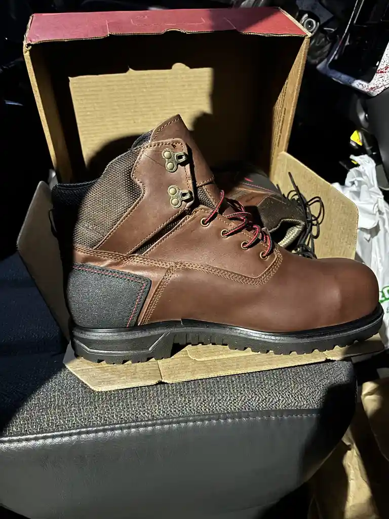A brown leather work boot with red laces sits inside its open cardboard box, resting on a car seat. The boot has a thick…