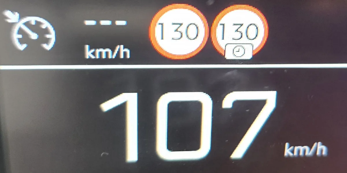 A car's digital dashboard shows the current speed as 107 km/h, with two orange circular warning icons above it displaying…