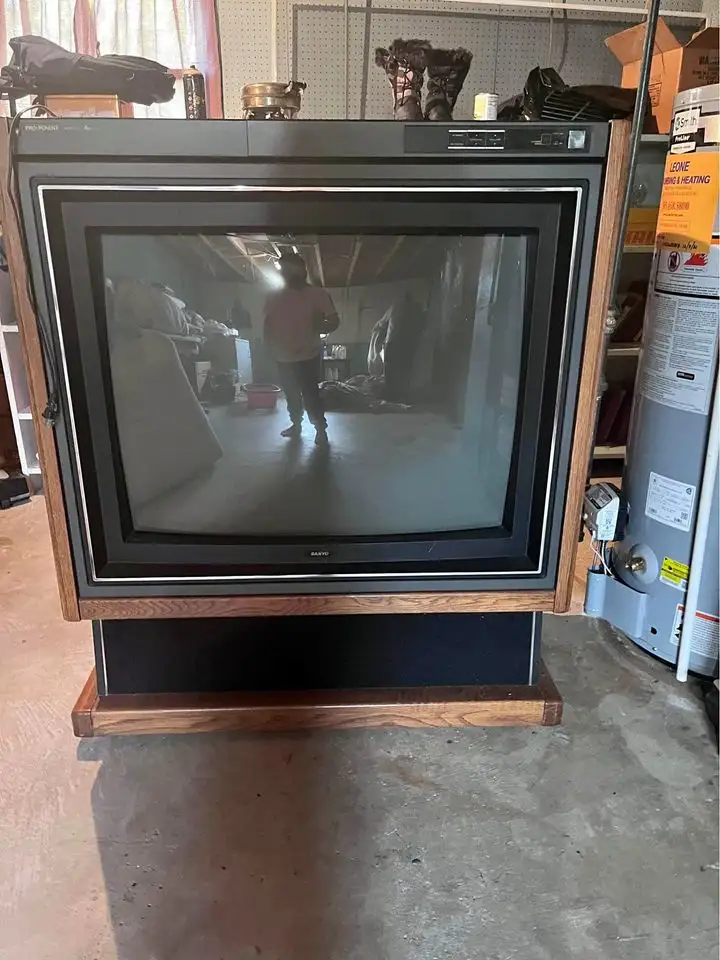 A vintage Sanyo CRT television with a wooden frame and stand sits in a cluttered garage or storage room. The TV screen is…
