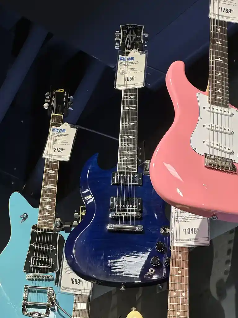 Three electric guitars are displayed in a store, each with a price tag: a light blue guitar priced at $999.99, a dark blue…