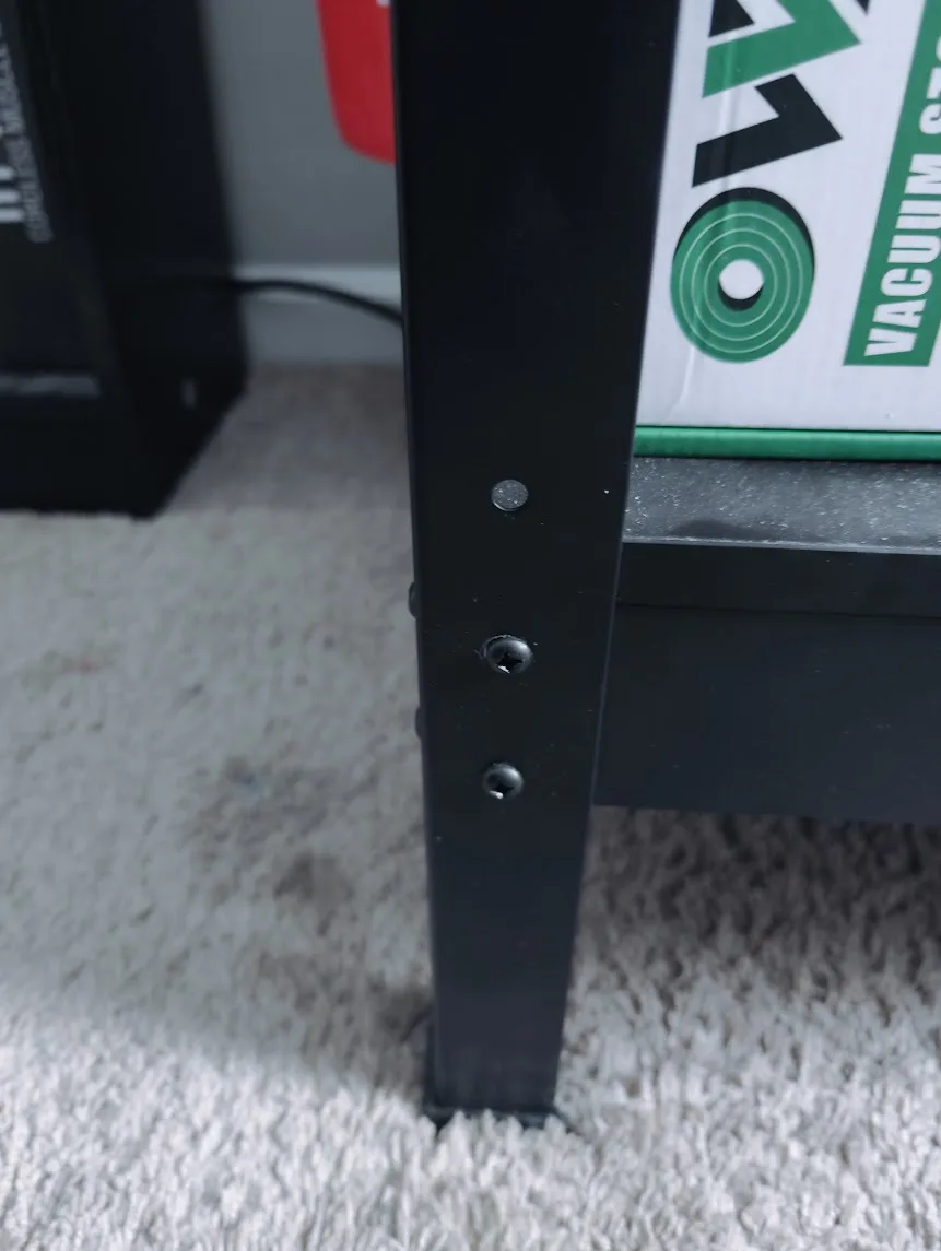 A close-up photo shows the leg of a black metal table or shelf with three visible screws, one of which is missing its head…