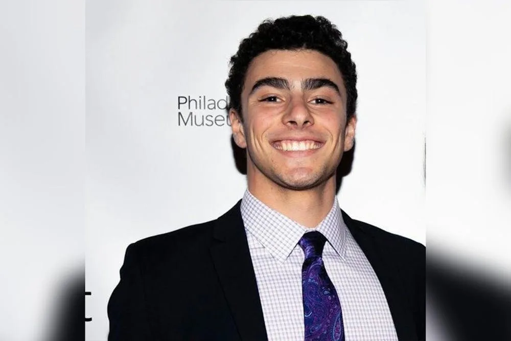 A young man with dark curly hair, wearing a black suit, light checkered shirt, and purple paisley tie, smiles broadly at the…