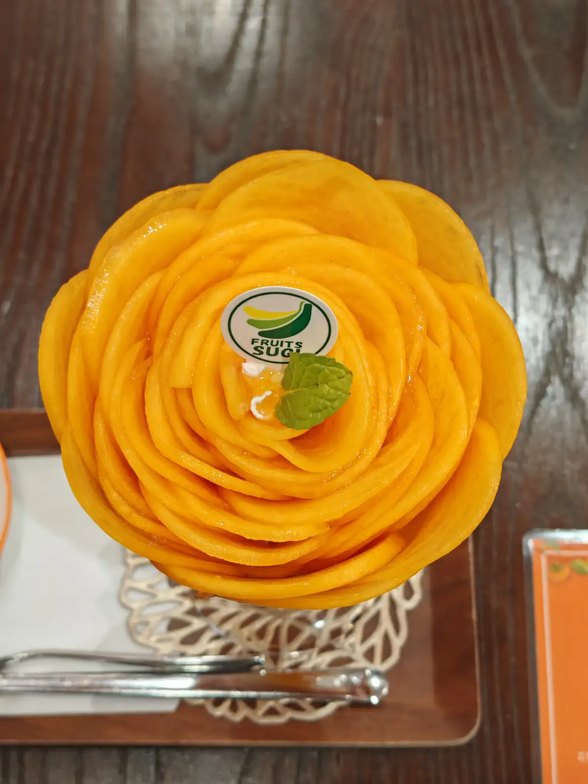 A top-down view of a dessert in a glass cup, shaped like a vibrant orange rose made from thinly sliced mango. At the center…