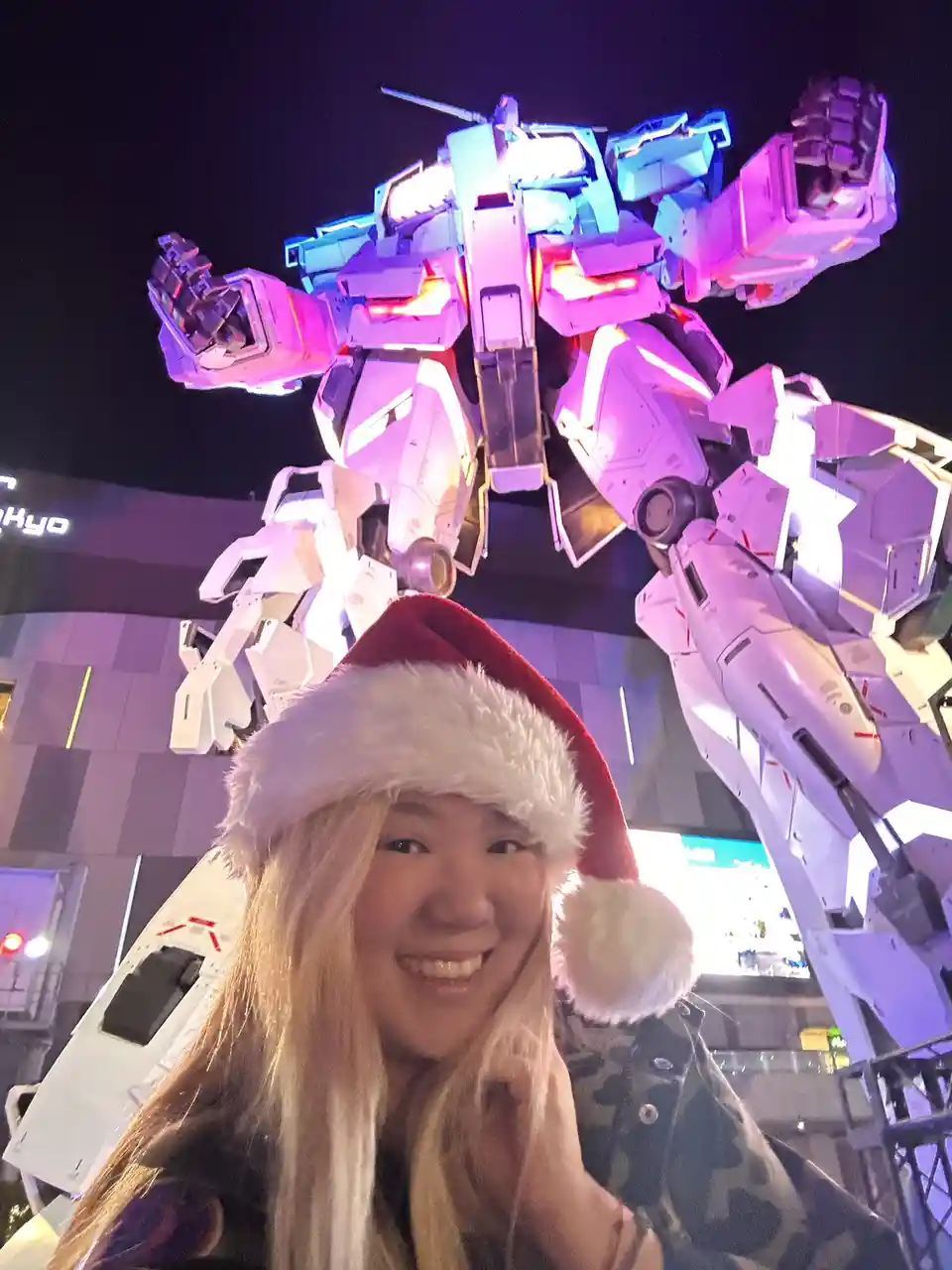A smiling person wearing a red and white Santa hat poses for a selfie in front of a massive, illuminated white and blue…