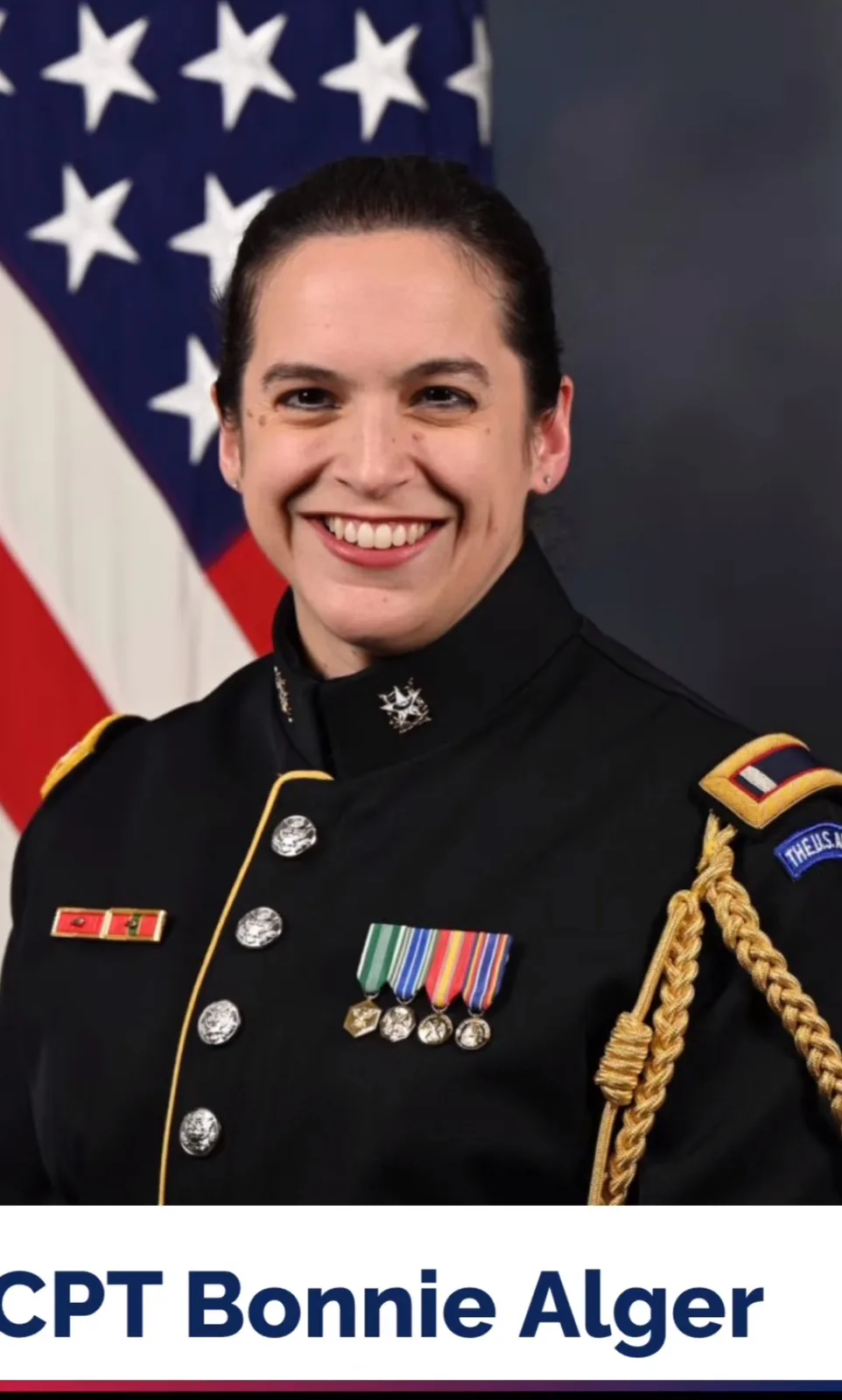 A smiling woman in a formal U.S. Army dress uniform with medals and a braided cord, standing in front of an American flag…