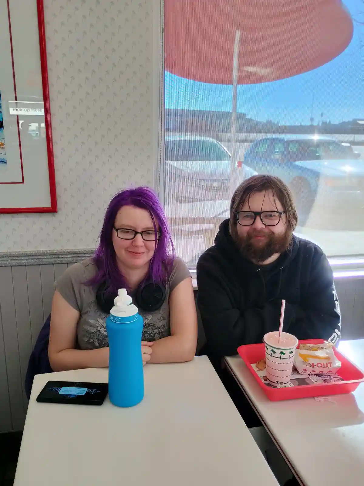 Two people sit at a fast-food restaurant table, one with purple hair and a blue water bottle, the other with a beard and a…