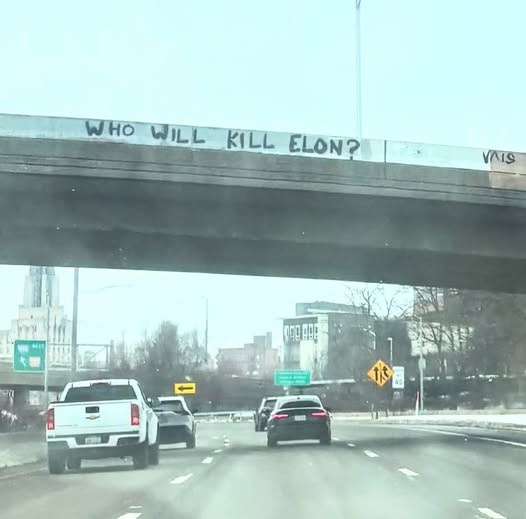 A photo taken from a car on a highway shows a concrete overpass with the phrase 'WHO WILL KILL ELON?' written in black…