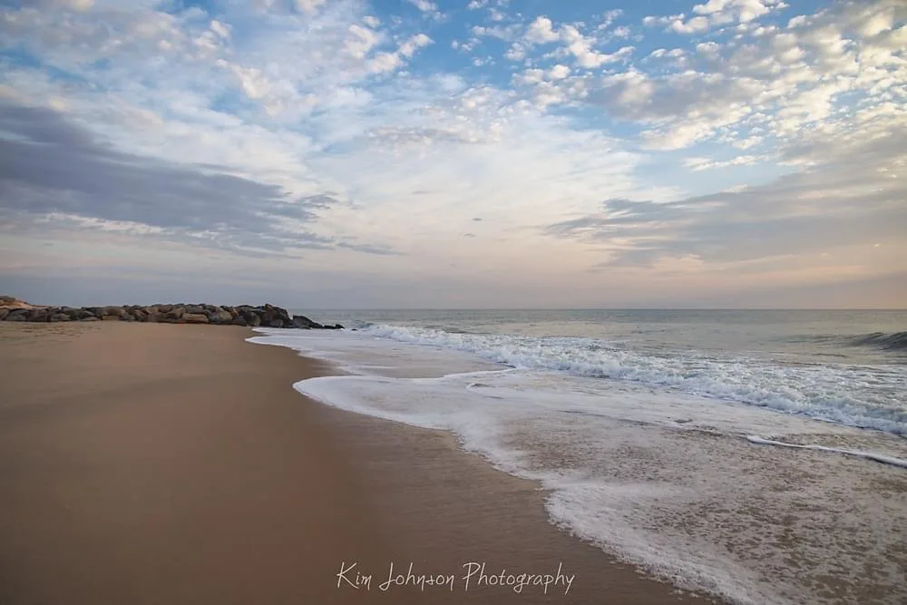 A serene beach scene with gentle waves rolling onto golden sand under a partly cloudy sky at sunrise or sunset. The photo is…