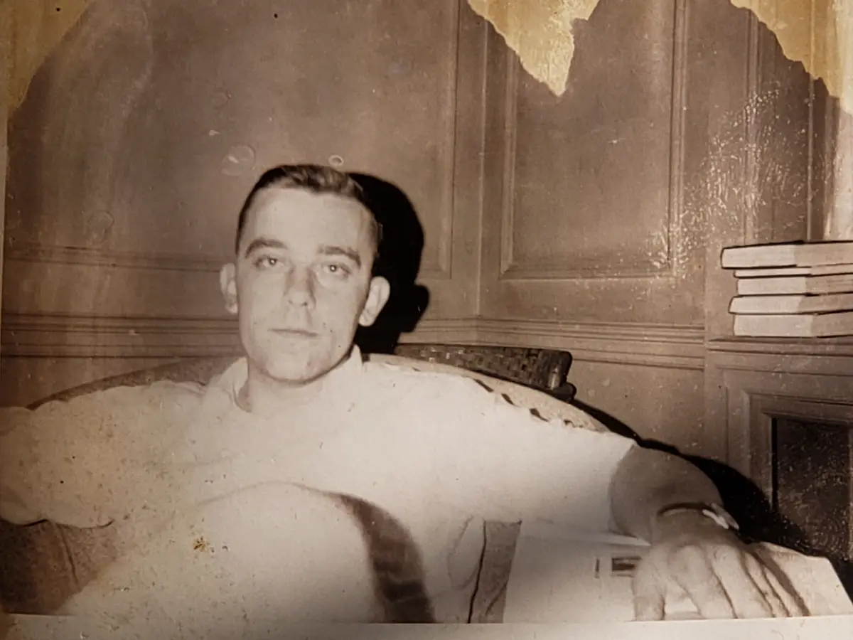 A sepia-toned, vintage photograph shows a man in a light-colored shirt sitting in a chair, looking directly at the camera…