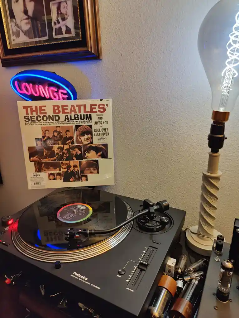 A Technics turntable plays a vinyl record, with a poster of 'The Beatles' Second Album' leaning against the wall above it…