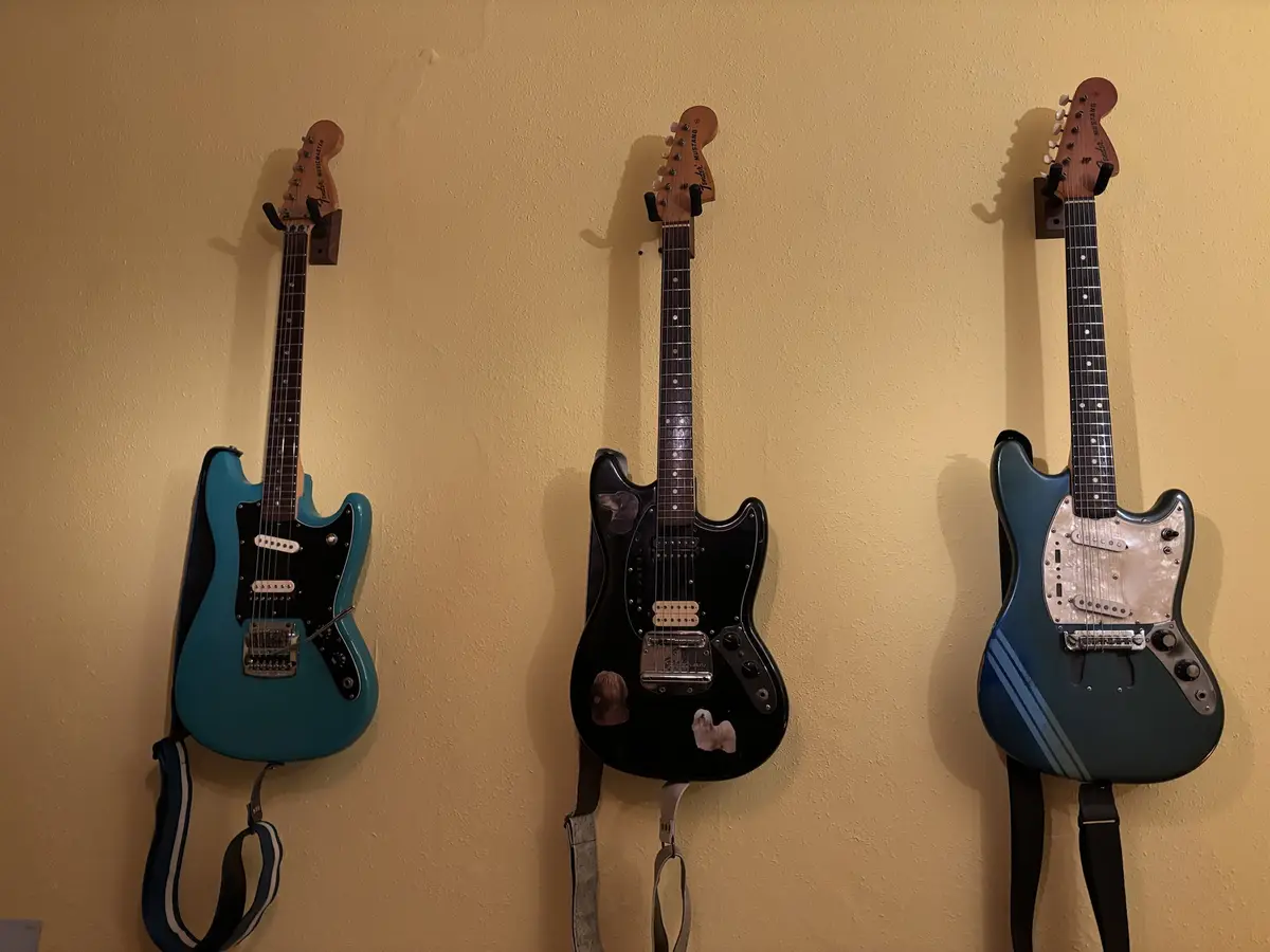 Three electric guitars hang vertically on a beige wall, each mounted on a wall bracket. The left guitar is a turquoise…