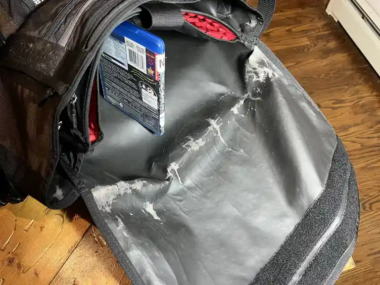 A black backpack with a heavily worn, torn, and stained interior sits open on a wooden floor, containing only a single blue…