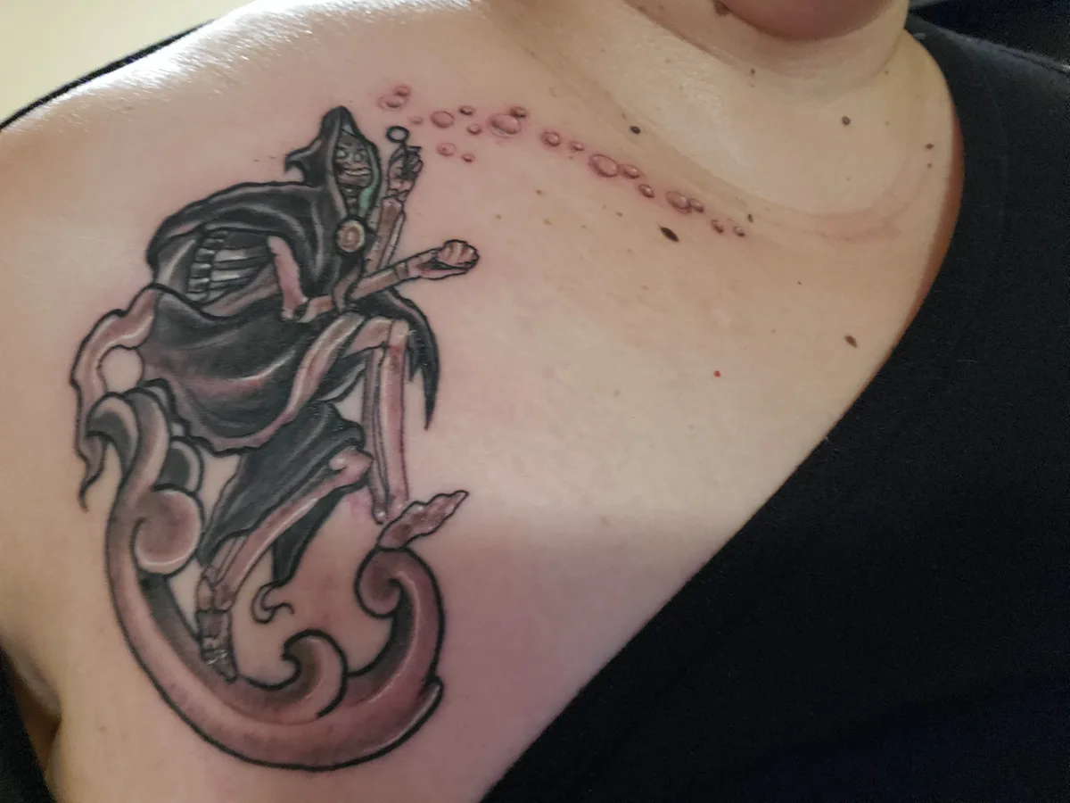 A close-up photo shows a freshly inked tattoo on a person's upper chest/shoulder area, depicting a stylized, hooded figure…