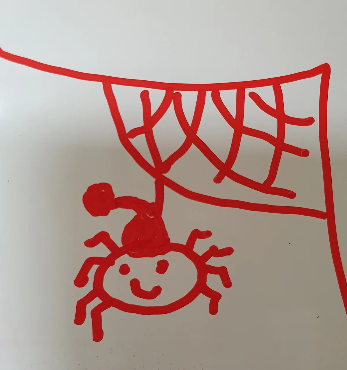 A child's red marker drawing on a white surface shows a stick figure with a round head and limbs, riding a smiling sun with…