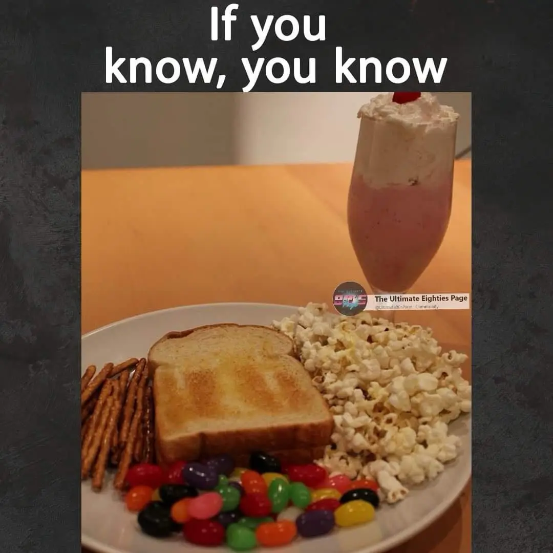 A plate of food including two slices of toast, pretzels, popcorn, and colorful jelly beans sits next to a tall glass of pink…