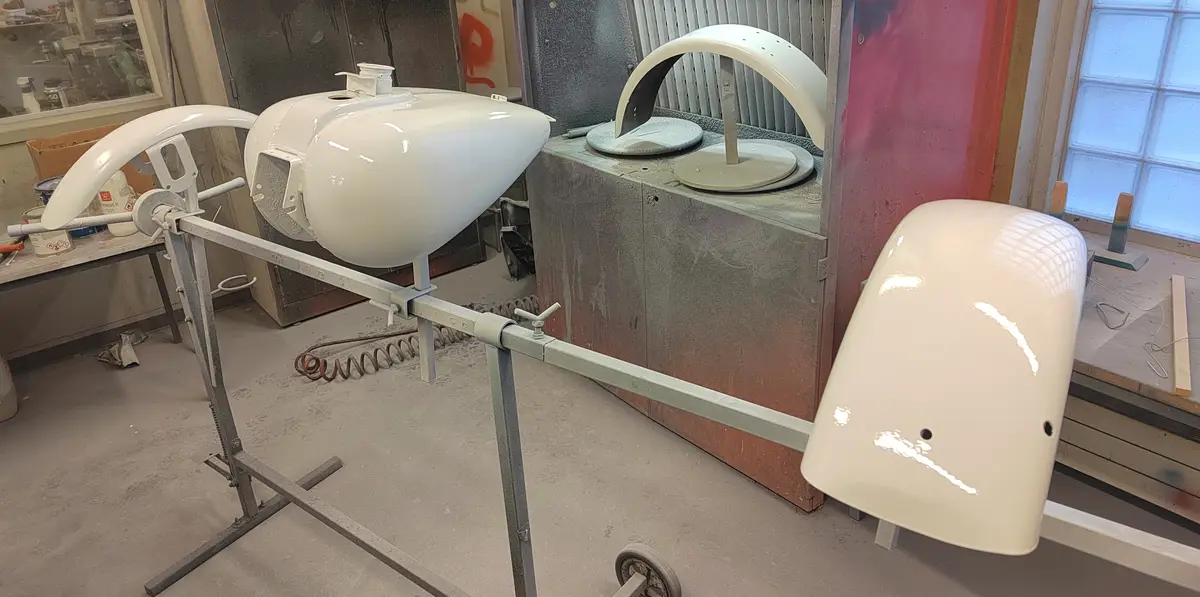 A workshop scene with several white-painted car body panels mounted on metal stands, including a fuel tank and fender-like…