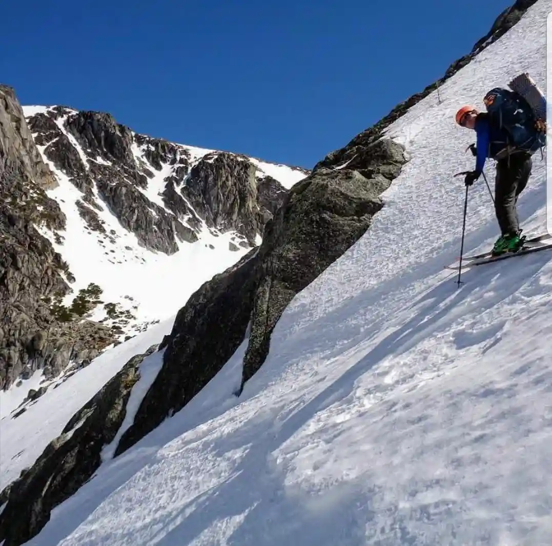 A skier in a blue jacket, orange helmet, and large backpack is ascending a steep, snow-covered mountain slope under a clear…