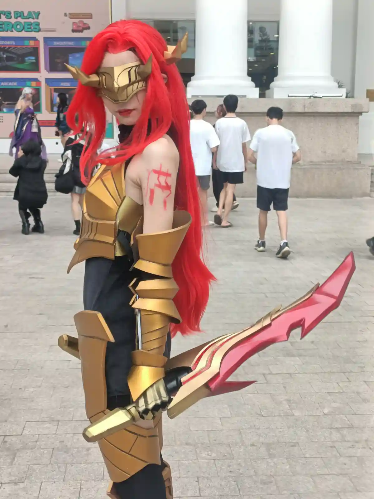 A person in a detailed cosplay of a video game character with long red hair, a gold horned mask, and gold armor holds a…