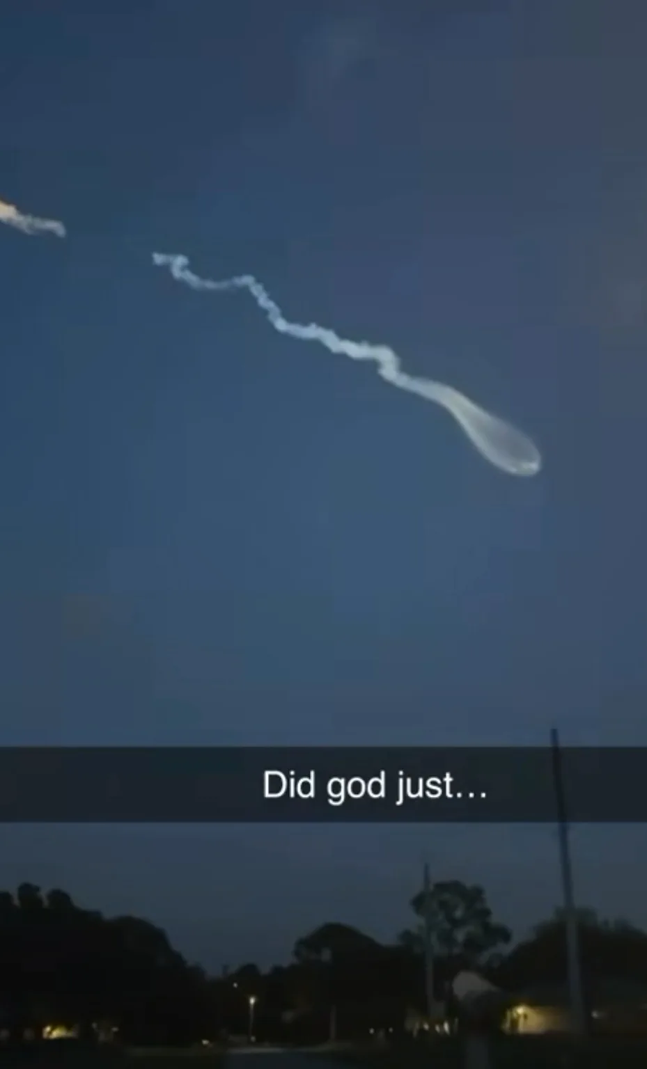 A dark sky at dusk shows a bright, elongated object with a trailing smoke-like vapor trail moving across the frame. The…